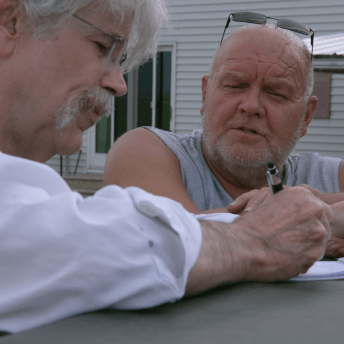 Still from Storm Lake. Art Cullen writes on paper, while other man watches him.