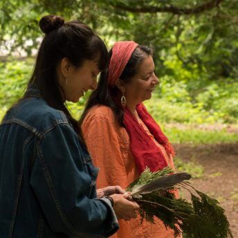 Two women, one young and the other one older, are standing in a forest. They smile.