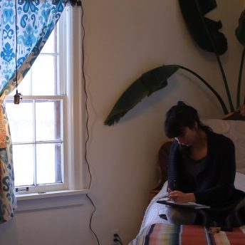 A young woman sitting in a bed writing in a notebook.