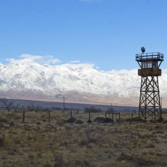 Still from Manzanar Diverted: When Water Becomes Dust. A watchtower stands in a field with a mountain range and blue skies in the background.