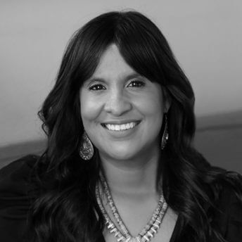A woman with dark skin, long, wavy black hair and black eyes sits on a bright red/orange sofa, her hands resting on the sofa's arm rest. She's smiling while looking into straight into the camera. She is wearing a long sleeve black dress with a V-neck,has on a turquoise necklace and matching earrings.