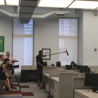 Production still of Angela Tucker looking into a camera monitor with her hand over her mouth and smiling. She is wearing glasses and has her hair in a bun. Standing next to her is a man wearing glasses and holding a notebook.