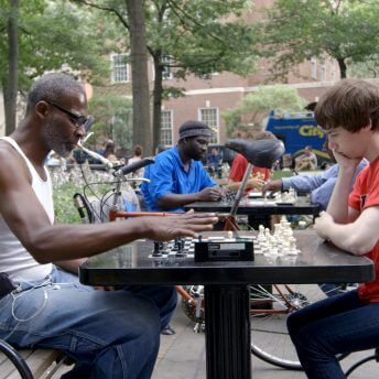 An older Black man and a younger white man study a chess board during a match in Washington Square Park, New York.
