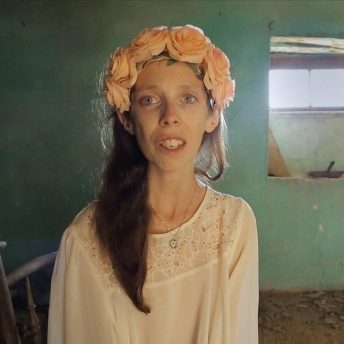 The photographer Lene Marie Fossen being interviewed at a shooting-site in Chios. She is looking directly into the camera lens, wearing a white dress and an apricot-colored hairpin of roses.