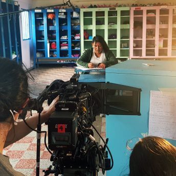 Sound person and camera person work in a scene where a young woman is sat on a school bench writing