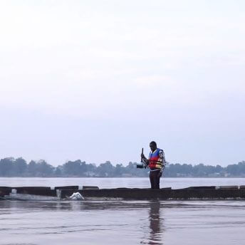 Eat Bitter cinematographer Orphe stands in the middle of a lean canoe while holding his camera towards the character Thomas, who is bent over using ropes to fix the canoe. On the right side of the canoe is Thomas' partner.