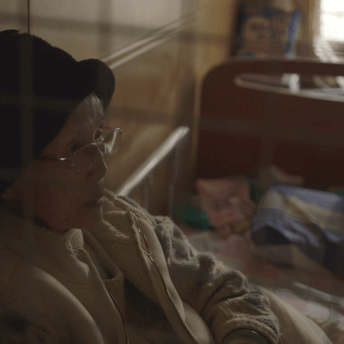 An older, Taiwanese woman with a black hat and glasses sits on her bed, behind a blurry grate in the foregound.
