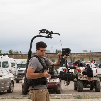 A cameraperson holds a camera harness, in the background cars and trucks are parked, there is a motorcycle with children looking at something