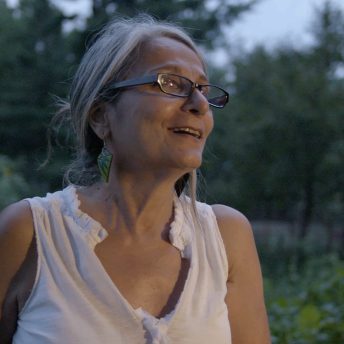 A middle-aged woman is standing in her backyard at dusk. Her face is illuminated and she is smiling and gazing out at something off-screen.