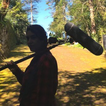 Director Indira Cato holding a boom microphone. Behind her, a dirt road and several trees on a rural landscape in Hidalgo, Mexico.