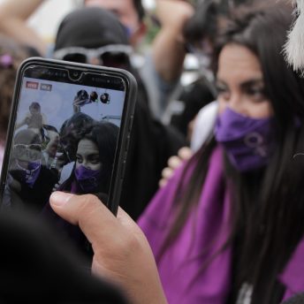 A phone recording Olimpia Coral and others outside the Chamber of Deputies before the Olimpia Law's approval. Olimpia wears a purple mask and a purple flag over her shoulders.