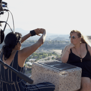 Chithra Jeyaram, a south Asian filmmaker, and Brandy Stein, a white woman, are sitting facing each other at a scenic outlook at magic hour. Chithra is facing away from the camera with her arms outstretched to block the sun while in conversation with Brandy, who is facing the camera. Brandy has sunglasses and her right elbow is resting on a plaque with the hand under her chin. In the foreground to the left, is a camera on a tripod, both out of focus.