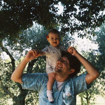 A man with a short sleeve shirt has a baby on his shoulders, he looks at her, the baby has a flower behind her ear and looks giggly at the camera
