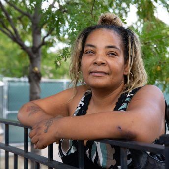 Medium shot of a woman resting her arms on a fence, wearing a tank top and looking up, in the background there are trees.