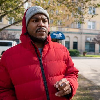 Medium shot of a man wearing a red puffed jacket and a gray beanie, in the background there is a tree and a building.