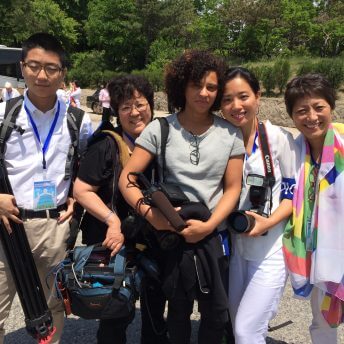 Medium shot of 5 people holding camera and audio equipment. Two of them, including director Deann Borshay Liem, are dressed in white. In the background there is a bus and people coming off of it.