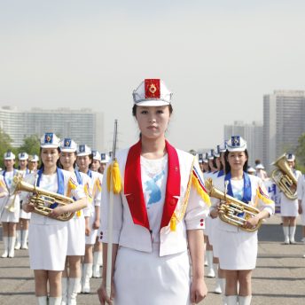 Medium shot of a Korean woman dressed in white, with the shape of Korea in her t-shirt, on the back there are more women in lines, dressed in white as well and some of them hold instruments.