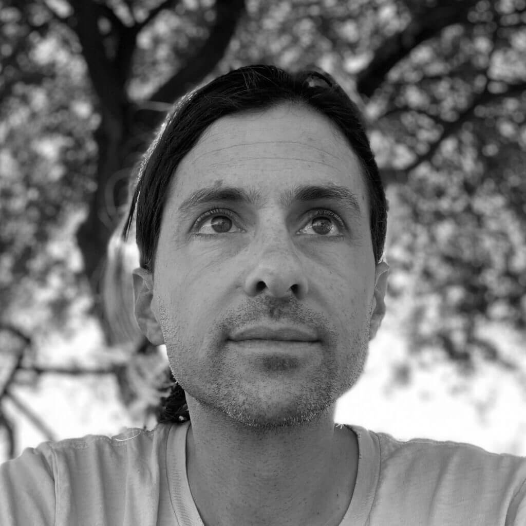 Portrait of Nick Kelso. He wears a shirt and looks away from the camera, in the background there is a tree. The portrait is taken in a low angle.