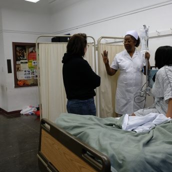 Director Emma Francis-Snyder talking to an actress dressed as a nurse, and an actress dressed as a patient sat on a bed
