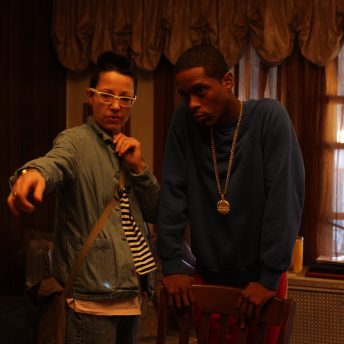 Production still of A Cops and Robbers Story. Ilinca Calugareanu and actor Corey Pegues Jr. standing in a low-lit room. They are looking off-camera. Corey is leaning forward with his hands on a dining room chair and Illinca has her arm extended as she points off-camera. There are windows behind them that reveal light shining through curtains.