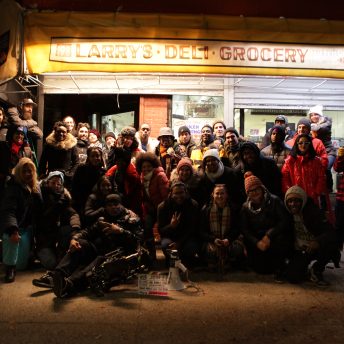 Production still of A Cops and Robbers Story. Crew, cast and documentary subjects are posing for a final photo at the end of the dramatizations shoot. They are outside at night and stand underneath a bodega's lit awning that reads "Larry's Deli Grocery."