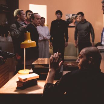 Still from Chuck Norris vs Communism. A Secret Police raid is shown and a man with his hands up looks to his left at a gun pointing at him. In the background and out of focus are a group looking on. (photographer Balla Zoltán)