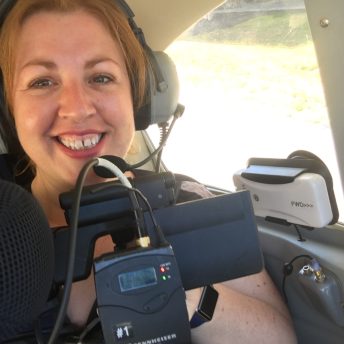 Production still of Jeanie Finlay looking directly at the camera and smiling. She is sitting in the back of a small Cessna plane, wearing headphones and holding a camera.