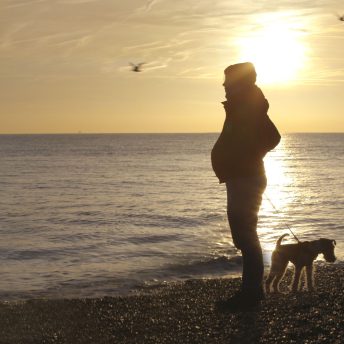 Still from Seahorse: The Dad Who Gave Birth. Freddy is standing on the seashore with his fox terrier dog. He is 9 months pregnant and is silhouetted by the sun.