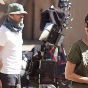 Production still of the 1491 TV series. Lisa Jackson standing in profile Taos Pueblo. She has short brown hair and is looking back and laughing. Some crew members stand near her and there is a camera behind her.
