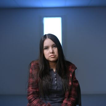 Still from Indictment: The Crimes of Shelly Chartier. Shelly Chartier sitting center-frame and looking directly at the camera. Behind her is a rectangular window and blue lighting on the wall.