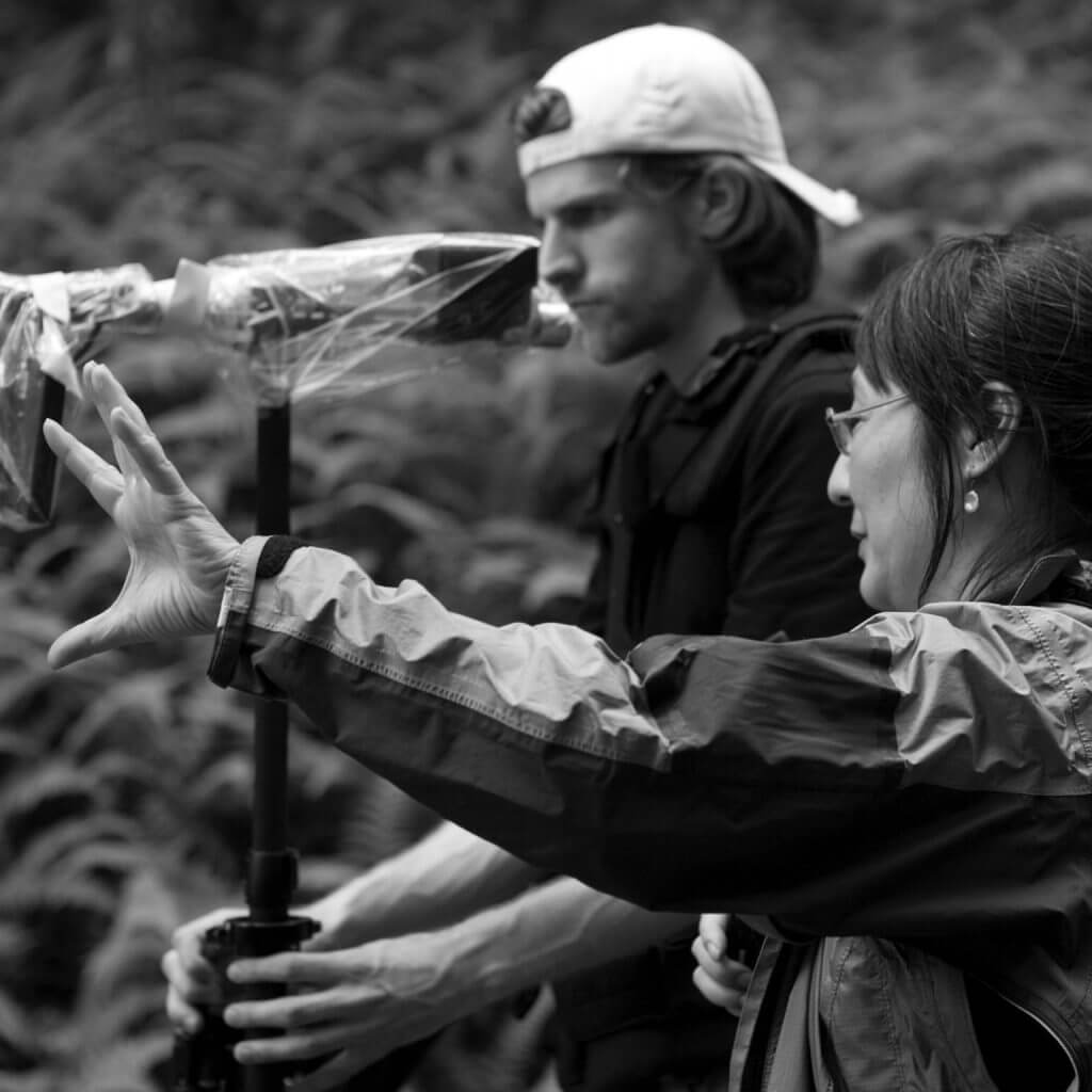 Production still of Rea Tajiri standing in a side profile to the camera. She is standing next to a man in baseball cap holding steadicam, and pointing to something in the distance. Black and white