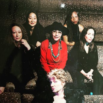 A group portrait of women directors directly facing the camera. Sitting in the center wearing a red shirt is Camille Billops. Moving clockwise, to her left is unknown person, above her is Rea Tajiri, to her right is Leslie Harris wearing a sleeveless black shirt, below her is another unknown person and sitting below her is Susan Seidelman wearing a black blouse.