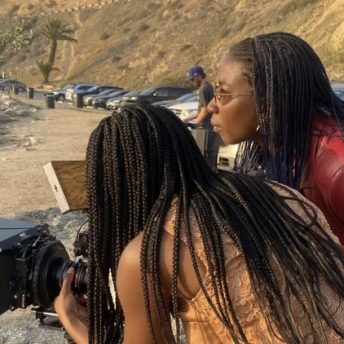 Production still of Sabaah Folayan standing in profile to the camera and wearing sunglasses. They are next to another woman with braids and a sleeveless blouse whose back is to the camera. They are both leaning forward and looking into a camera, which is pointed offscreen.