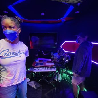 Production still from Look at Me. Sabaah Folayan standing in front of a piano keyboard in a music studio that is lit with blue LEDs. A man stands next to them with his back to the camera, looking into a large camera.