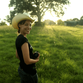 Sonia Kennebeck looking directly at the camera. She is standing in a green field with a tree in the background and is holding a photo lens.