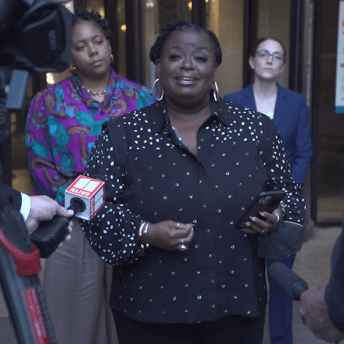 Production still of After Roe. Monica Simpson, Executive Director of SisterSong, speaking at a press conference after the first day of the SisterSong v Georgia trial. She is directly facing the camera and is wearing hoop earrings and a black blouse with gold polka dots.