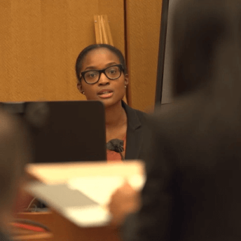 Production still of After Roe. Dr. Whitney Rice sitting and testifying in court during the SisterSong v Georgia trial. She is wearing glasses and is speaking towards a blurred woman in the foreground, who is at a podium and has her back to the camera.
