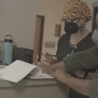 Production still of After Roe. Two women stand in profile to the camera. Both are masked and leaning over a desk, examining forms. Various items lay on the desk beside them, including hand sanitizer, a thermos and a water bottle.