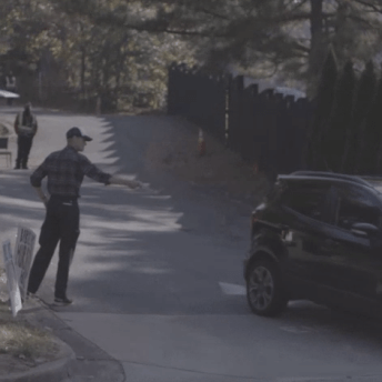 Production still of After Roe. A man in a hat and plaid shirt stands next to a group of protest signs. He has his arm extended towards a car that is leaving the Feminist Women's Health Center.