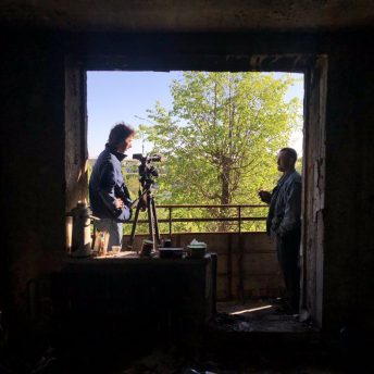 Production still of Displaced. The camera is placed in profile to two men framed in the background by a doorway. To the left is Volodymyr Usyk, Director of Photography, talking with a local resident. Trees are additionally seen in the background.