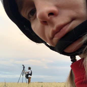Production still of Displaced. Olha Zhurba, director of Displaced, is taking a photo of herself and is shown in a close-up. In the background is Volodymyr Usik, Director of Photography, who stands in profile and looks up at a camera. They are close to the frontline, and are both wearing flat jackets and helmets.
