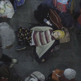 Still from Displaced. A girl is lying on family belongings at the train station. She looks directly up at the camera as other citizens walk past her.
