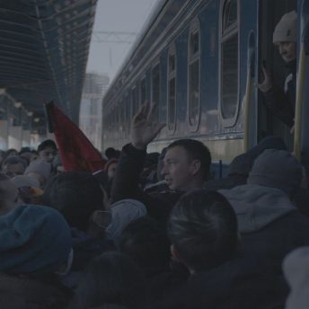 Still from Displaced. A main railway station in Kyiv, Ukraine during the first days of war. A crowd of people, who are attempting to get on an overcrowded train and evacuate the city, fills the photo. A man in profile to the camera extending his arm can be seen in the middle.