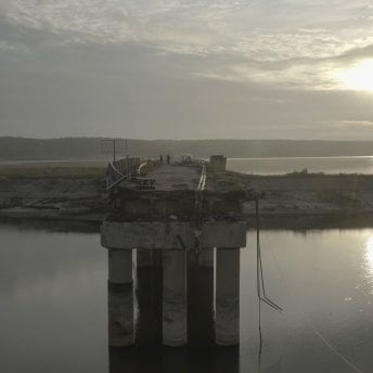 Production still of Intercepted. A destroyed bridge near the Kharkiv region is shown. Various rubble from the bridge hangs above a body of water. In the background, are two distant people standing on the bridge as the sun is setting.