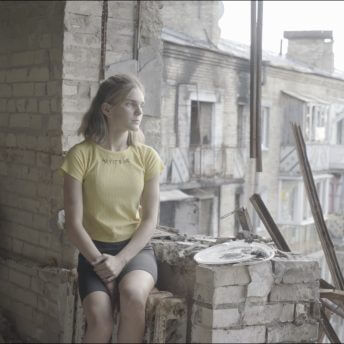Production still of Intercepted. A young teenage girl in a yellow shirt is sitting in profile to the camera. She is on the edge of her former apartment's window. Surrounding her is rubble, the result of a recent bombing. In the background, the exterior of another bombed building can be seen.