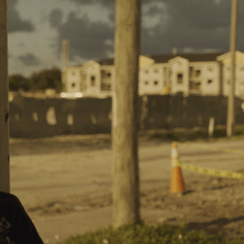 Still from Razing Liberty Square. A young boy is sitting in a side profile to the camera. He is smiling off camera to his left. Behind him are new buildings are being constructed, which are out of focus.