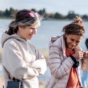 Production still of Sol in the Garden. Co-directors Emily Cohen Ibañez are looking towards the ground and laughing. Emily is on the left and has grey and dark brown hair and is wearing a beige sweatshirt. Débora is on the right and has dark brown, curly hair and is wearing a light purple puffer jacket. The ocean can be seen out of focus in the background.