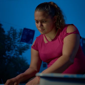 Still from Sol in the Garden. A woman with a hot pink shirt is looking down against a backdrop of blue sky at dawn.