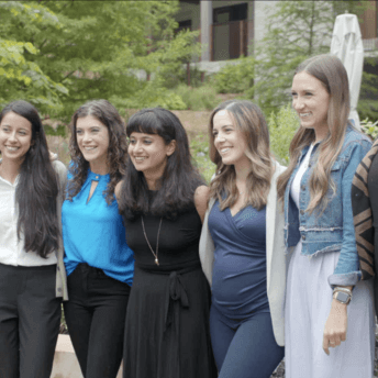 Production still of The Untitled 19th* News Film. News reporters and staff gather together in a casual, smiling group photo. They are facing a camera to the left. Behind them are trees and greenery, which are out of focus.