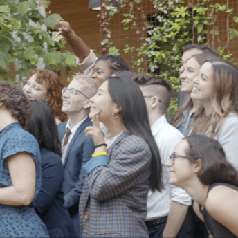 Production still of The Untitled 19th* News Film. News reporters and staff line up for a formal group photo. They are in a side profile to the camera and are smiling towards the left of frame.
